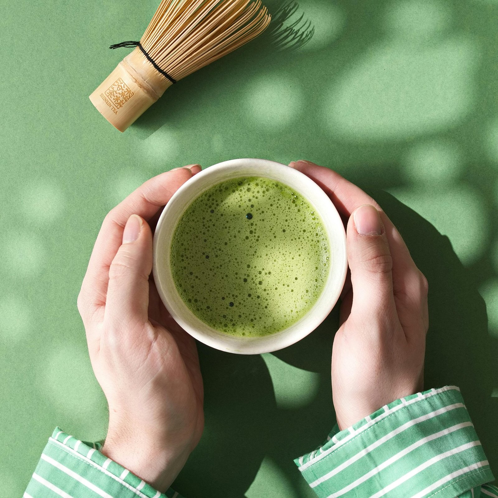Top view of hands holding a matcha bowl and bamboo whisk on green background.